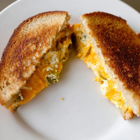 A close-up of Jalapeño Popper Grilled Cheese shows crispy buttery sourdough oozing with spicy, creamy cheese filling, perfect alongside a bowl of tomato soup.