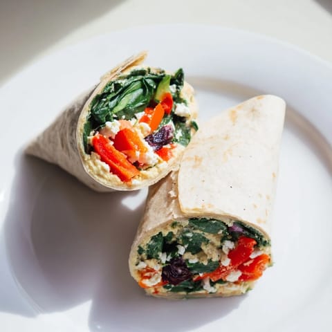 The sliced Mediterranean Veggie Wrap reveals layers of tangy feta cheese, bright red peppers, and fresh greens, served on a rustic wooden cutting board.  
